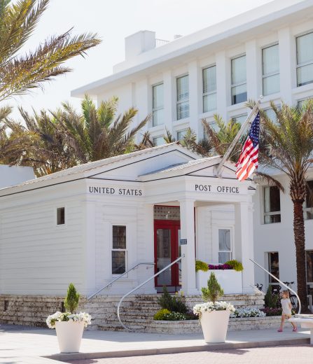 Small white United States Post Office building surrounded by palm trees, American flag, and modern white architecture.