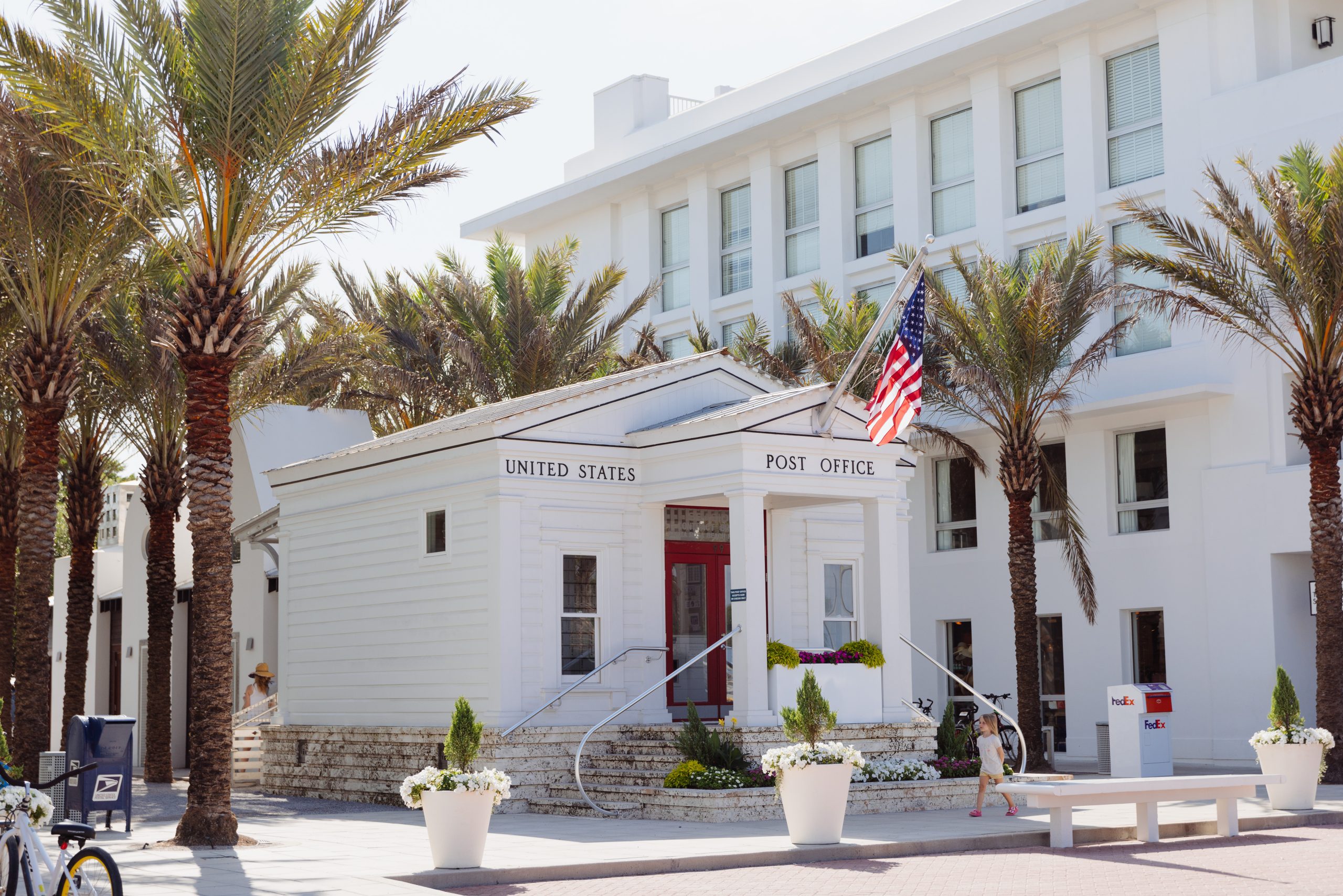 Small white United States Post Office building surrounded by palm trees, American flag, and modern white architecture.