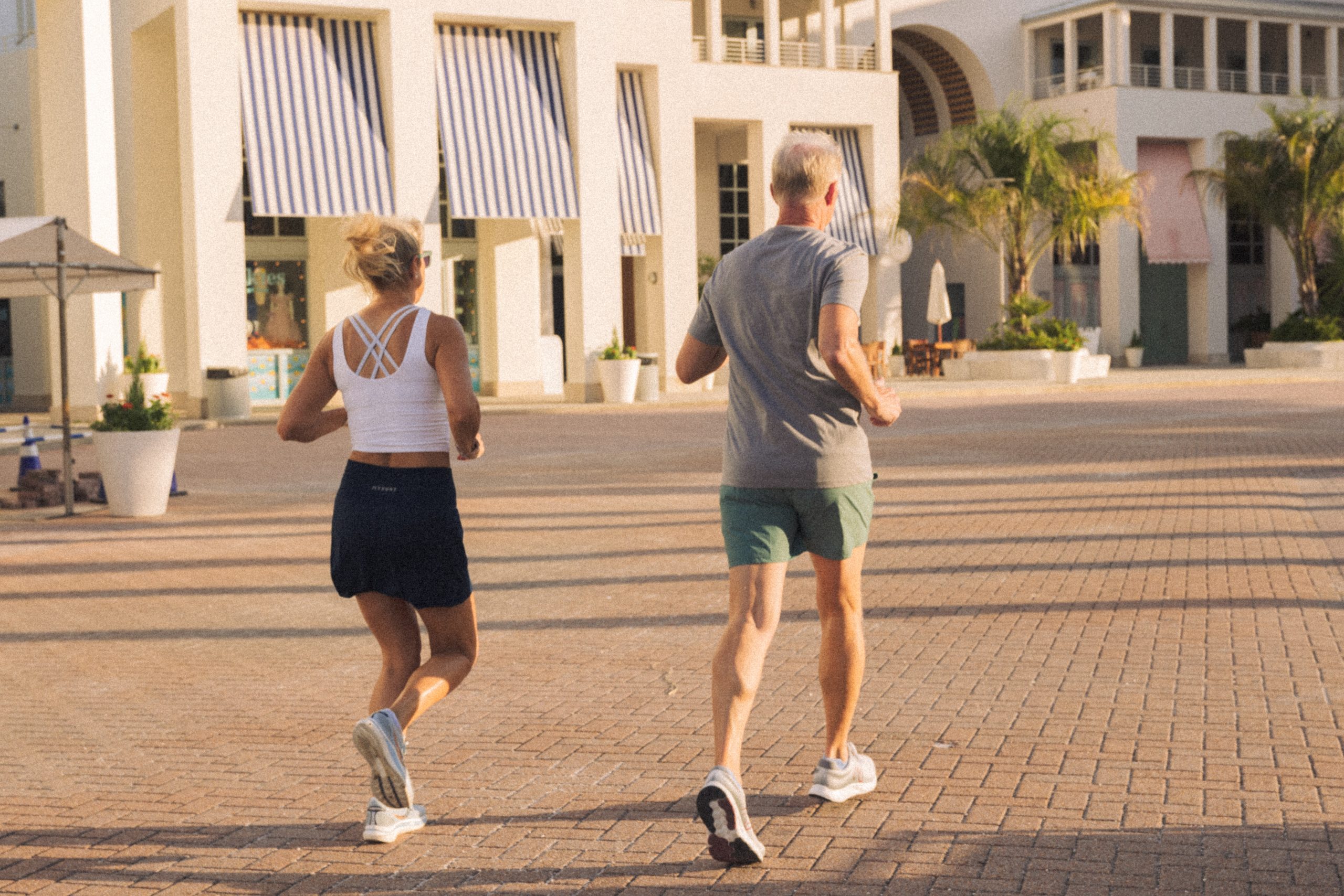Man and woman jogging together on a sunny morning near modern white buildings with striped awnings.