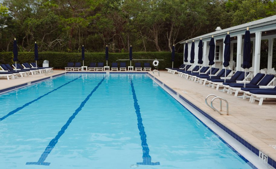 Empty swimming pool with blue lounge chairs and umbrellas surrounded by trees on a calm day.