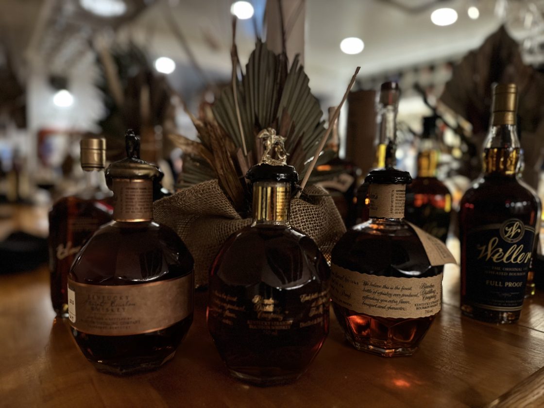 Close-up of whiskey bottles, including Weller and Blanton’s, displayed on a wooden bar counter.