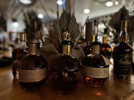Close-up of whiskey bottles, including Weller and Blanton’s, displayed on a wooden bar counter.