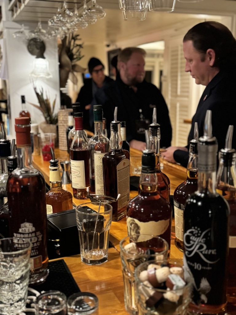 Whiskey bottles lined up on a wooden bar as a bartender serves drinks in a warmly lit lounge