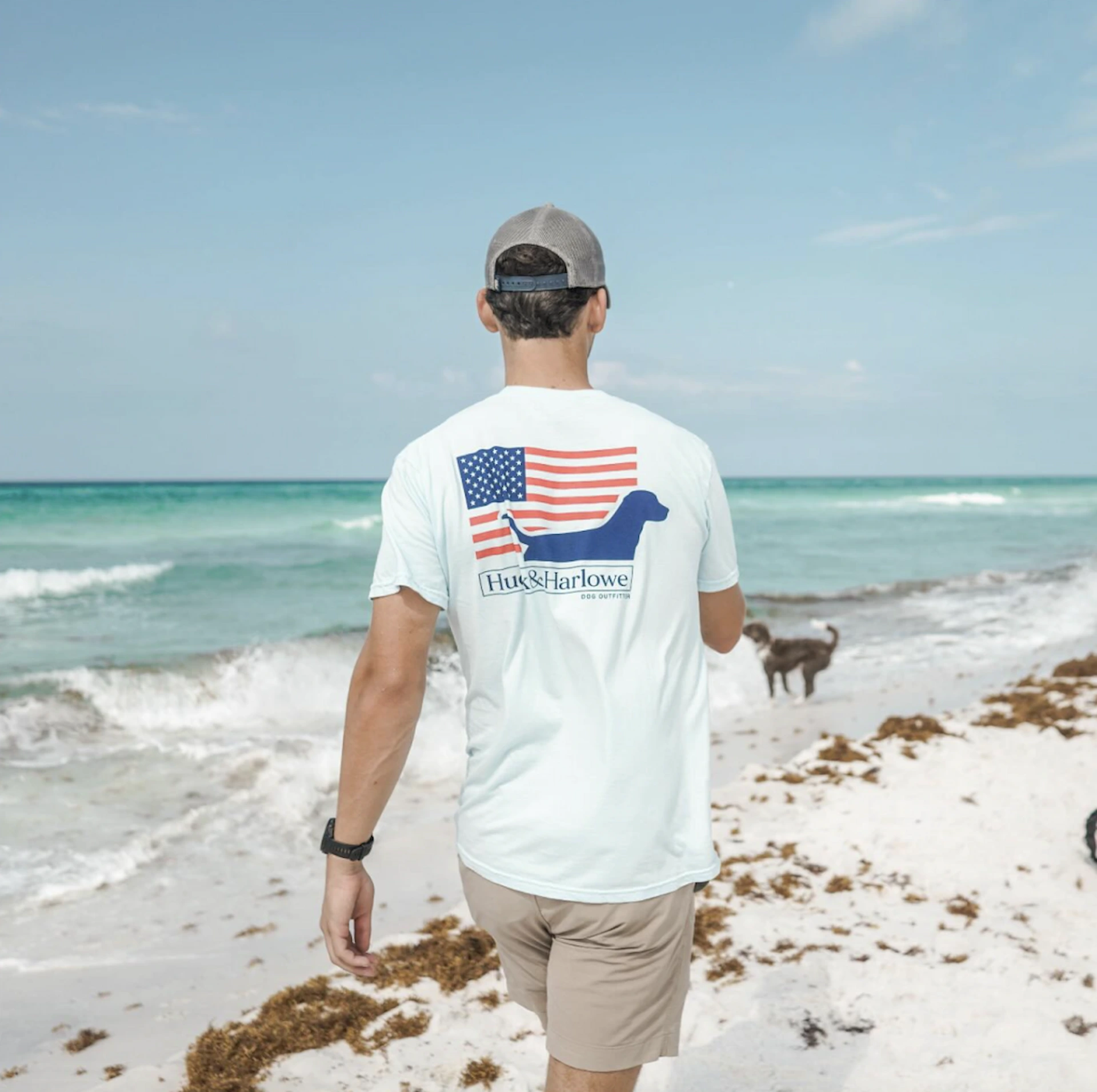 Person walking on a beach wearing a graphic T‑shirt with a dog design while a dog stands near the shoreline