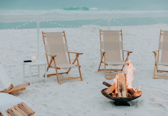 Campfire in a beach firepit surrounded by empty canvas chairs on white sand.