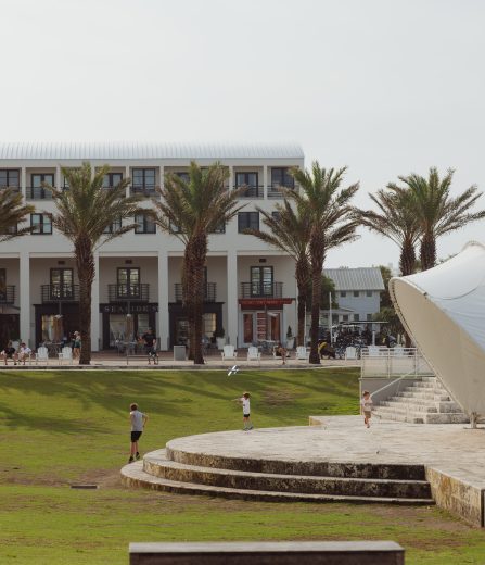 Children play on grassy amphitheater beside white pavilion, palm trees, and a modern building in background.