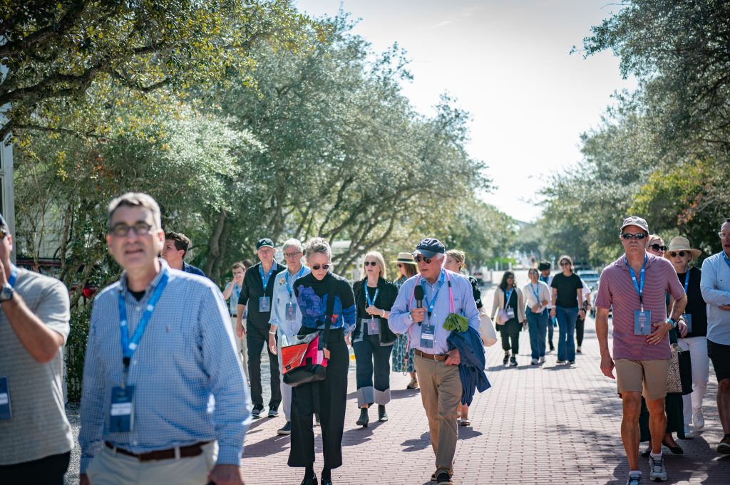 Groups of people wearing badges walk outdoors on brick path, surrounded by trees and bright daylight.