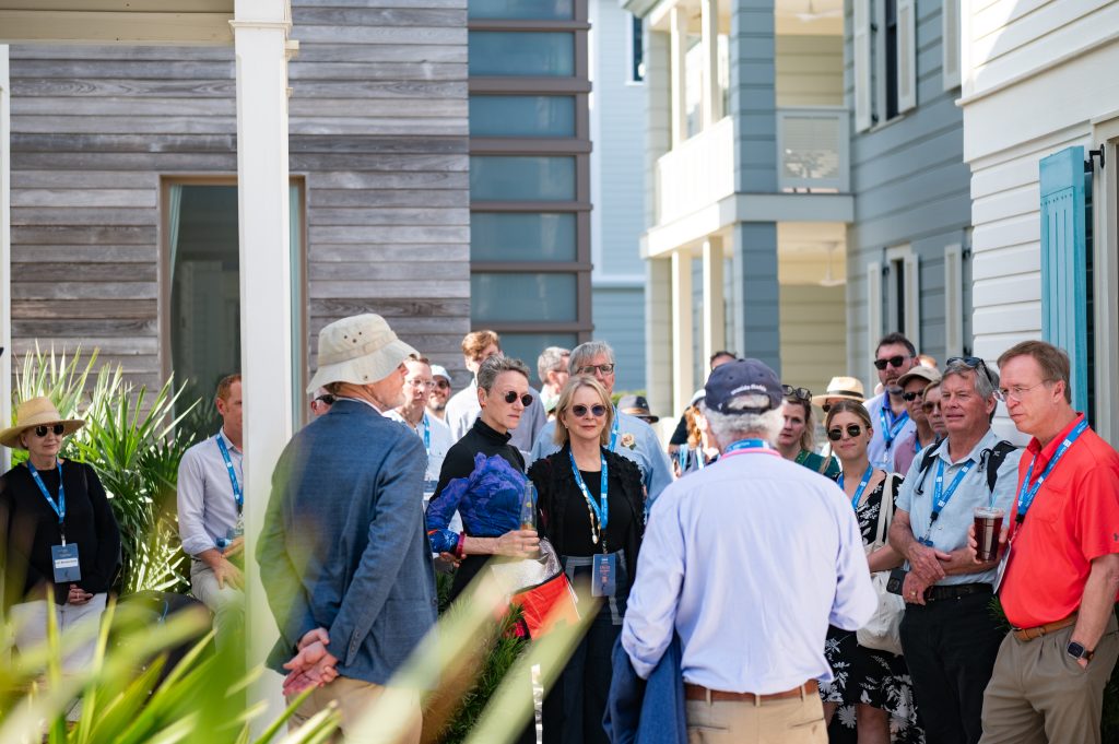 A group of people gathered outdoors, wearing event lanyards, chatting near modern building during daytime.