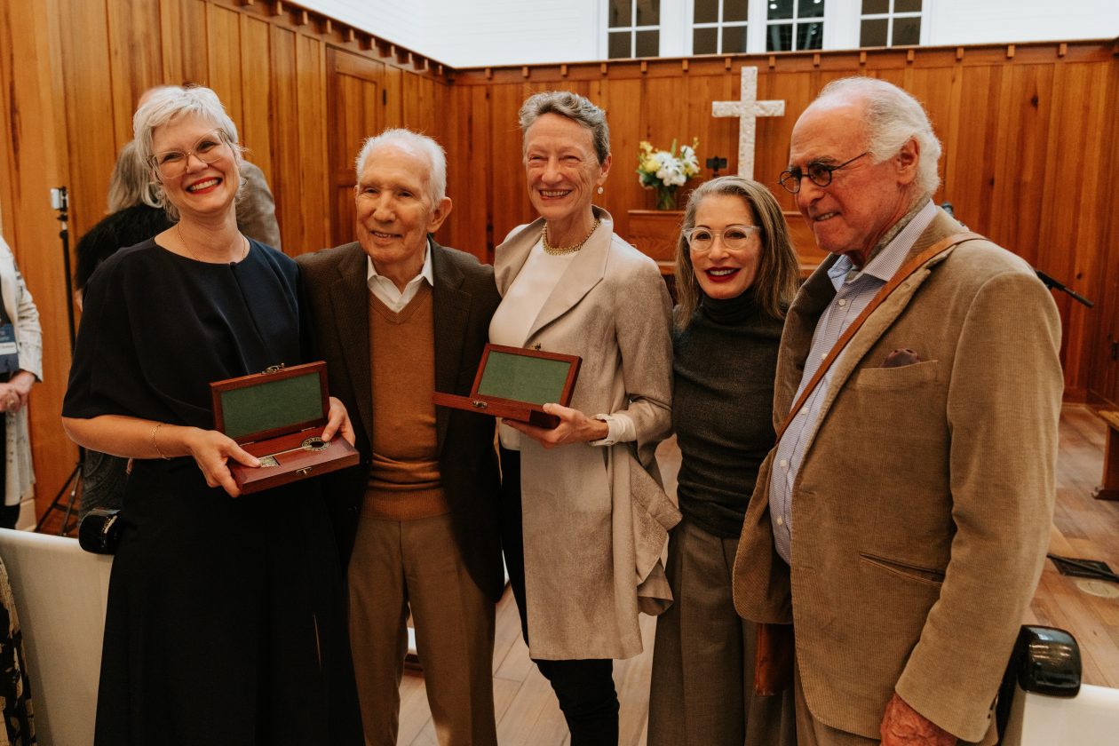 Five smiling adults in formal attire pose indoors holding award boxes in a wooden-paneled room.