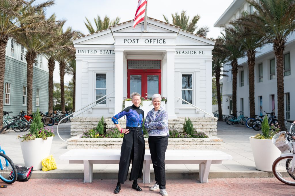 Two women stand smiling in front of the Seaside, Florida post office surrounded by palm trees and bicycles.