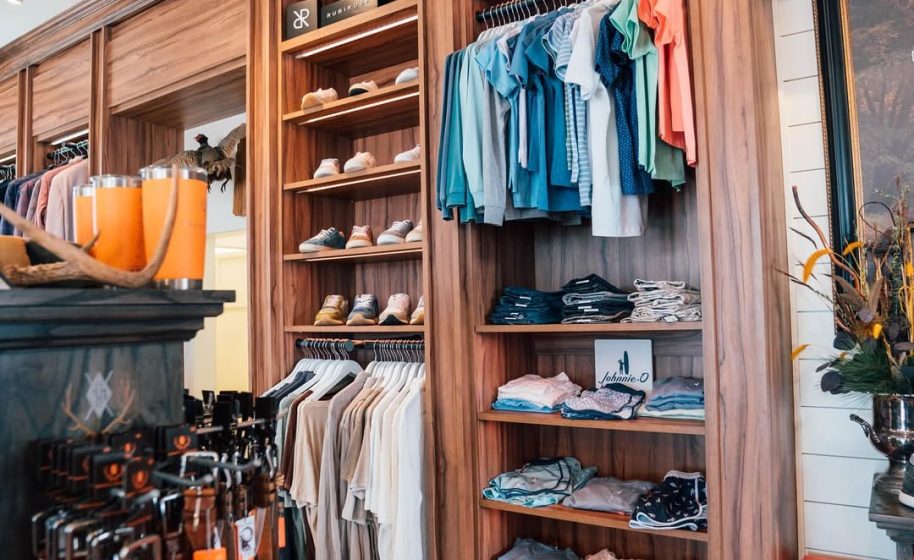Neatly organized clothing store with wooden shelves displaying folded shirts, hanging garments, shoes, and accessories.