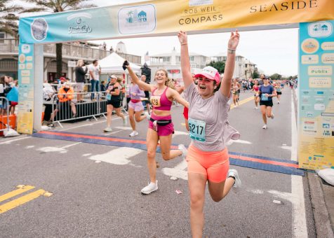 Runner in pink shorts and cap crosses colorful race finish line with arms raised, celebrating.