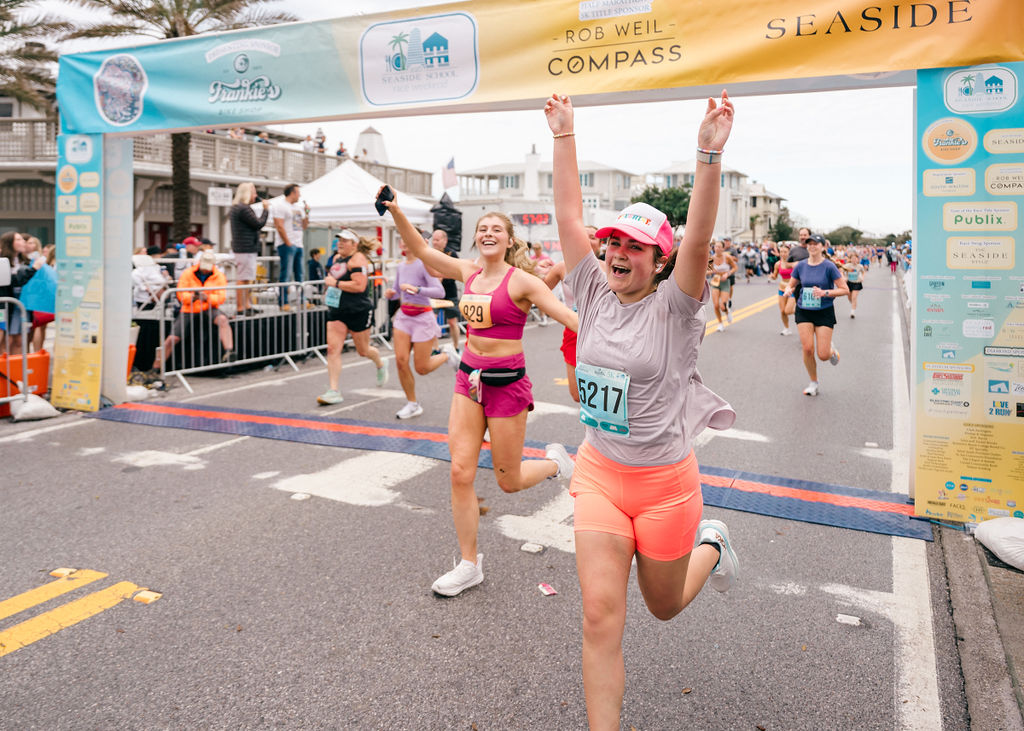 Runner in pink shorts and cap crosses colorful race finish line with arms raised, celebrating.