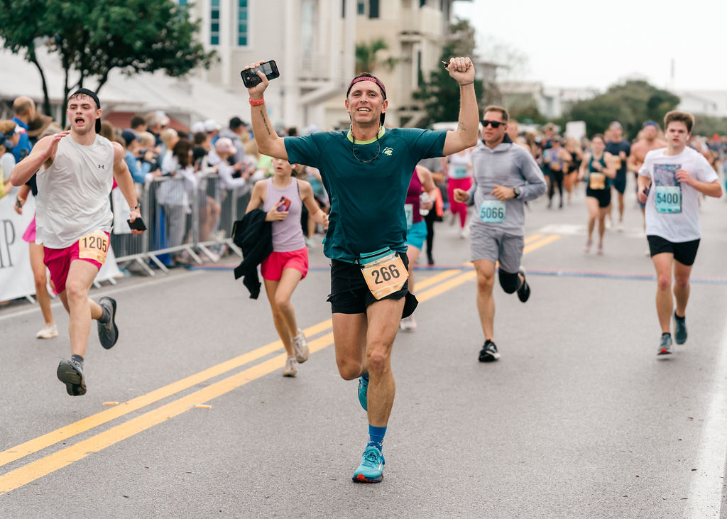 Runner celebrating with raised arms while crossing crowded road race finish line.