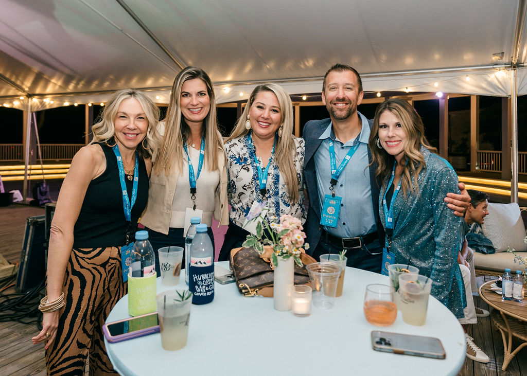 Smiling group at outdoor evening event gathered around cocktail table under a tent.
