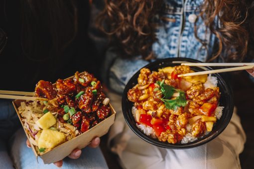 Two friends eating colorful Asian-style chicken rice bowls with chopsticks.