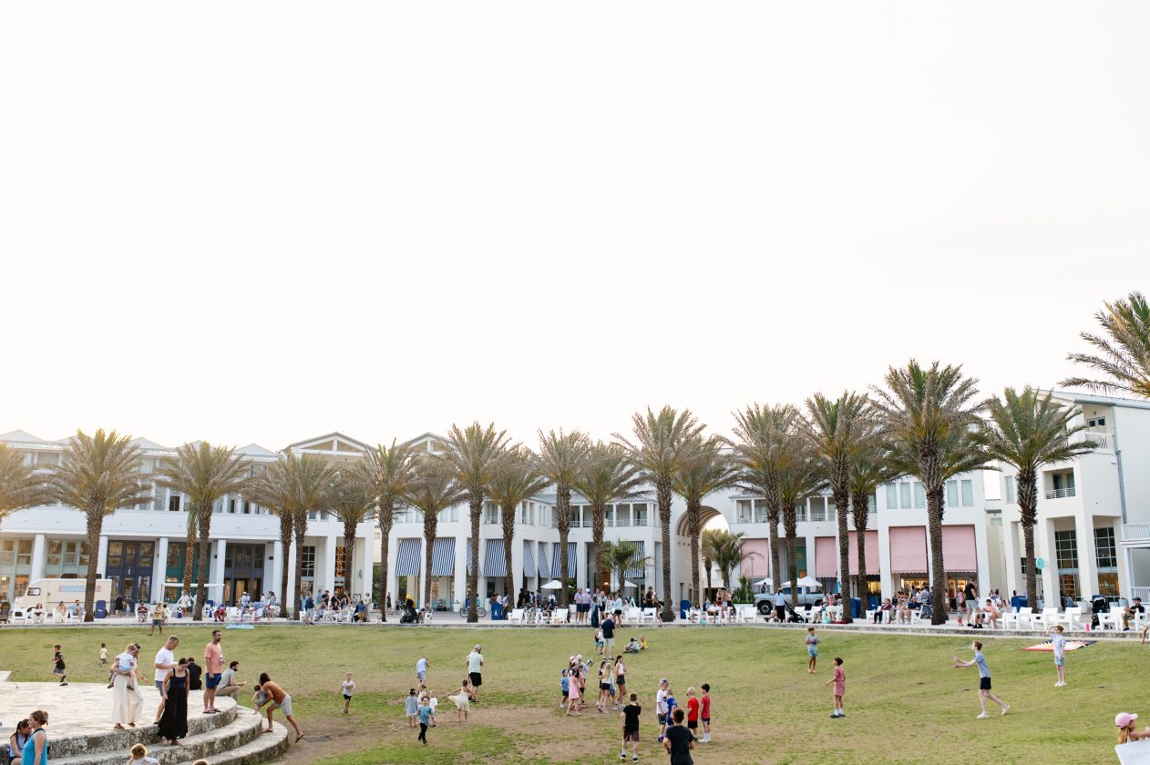 Families and children gathering on a green lawn in front of palm-lined seaside shops.