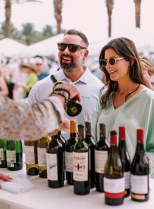 Two people wearing sunglasses at an outdoor wine tasting table with assorted bottles and a server pouring wine.