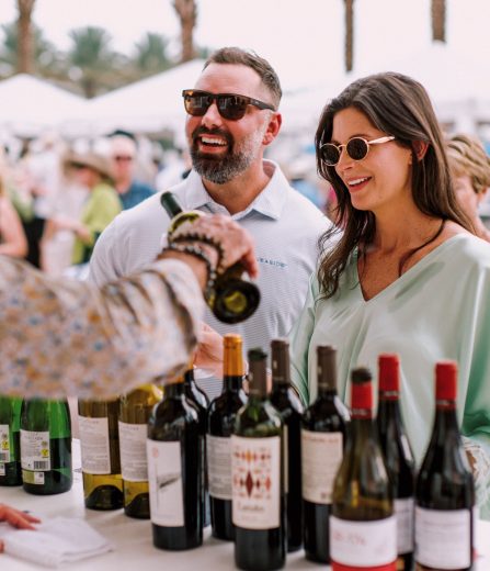 Two people wearing sunglasses at an outdoor wine tasting table with assorted bottles and a server pouring wine.