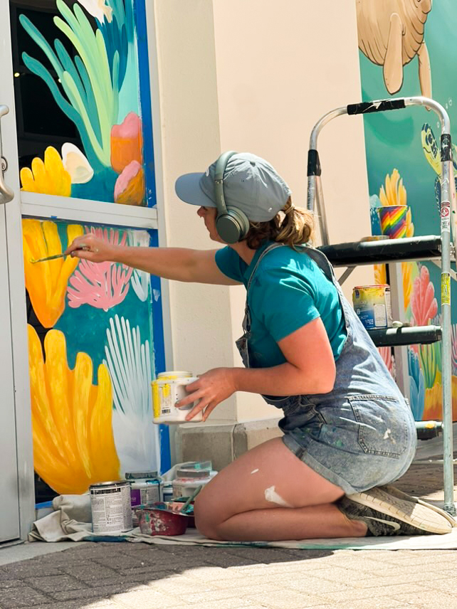 Artist wearing headphones paints a colorful underwater-themed mural with coral and sea plants on an exterior wall.
