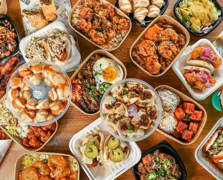 Assorted Asian-inspired dishes including rice bowls, dumplings, and bao buns arranged on a wooden table.