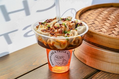 A plastic cup filled with dumplings topped with sauce and green onions sits beside a bamboo steamer.