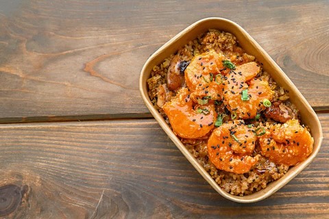 Takeout container filled with shrimp over fried rice, garnished with sesame seeds and chopped scallions.
