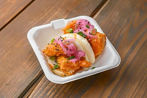 Two bao buns filled with crispy chicken, pickled onions, scallions, and sesame seeds in a container.