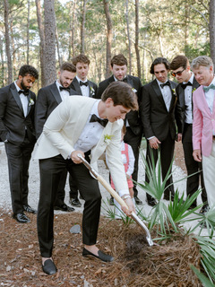 Groom pours water onto a plant as groomsmen watch during an outdoor wedding ceremony in a wooded setting.