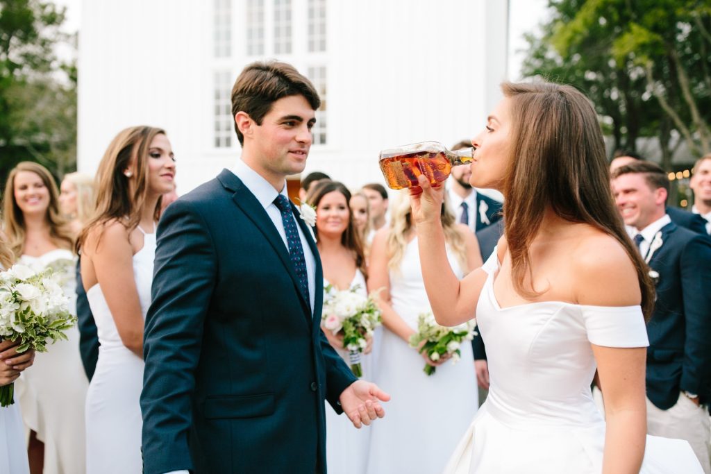 Bride drinks from a glass bottle as groom and wedding party watch during an outdoor wedding celebration