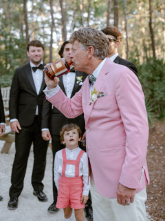 Man in a pink suit drinks from a flask while a child in suspenders and wedding guests look on outdoors.