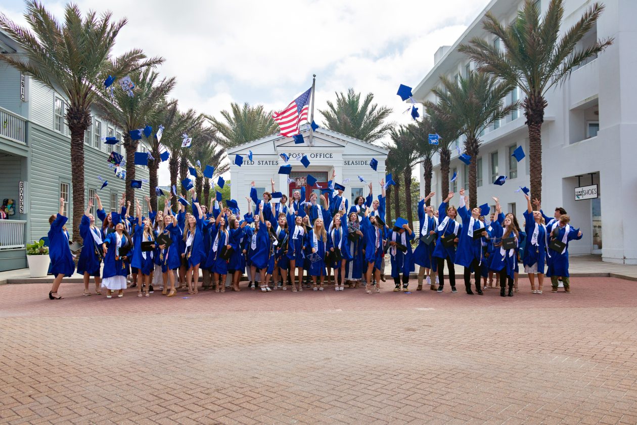 Graduates in blue caps and gowns celebrating outdoors with caps tossed in the air.