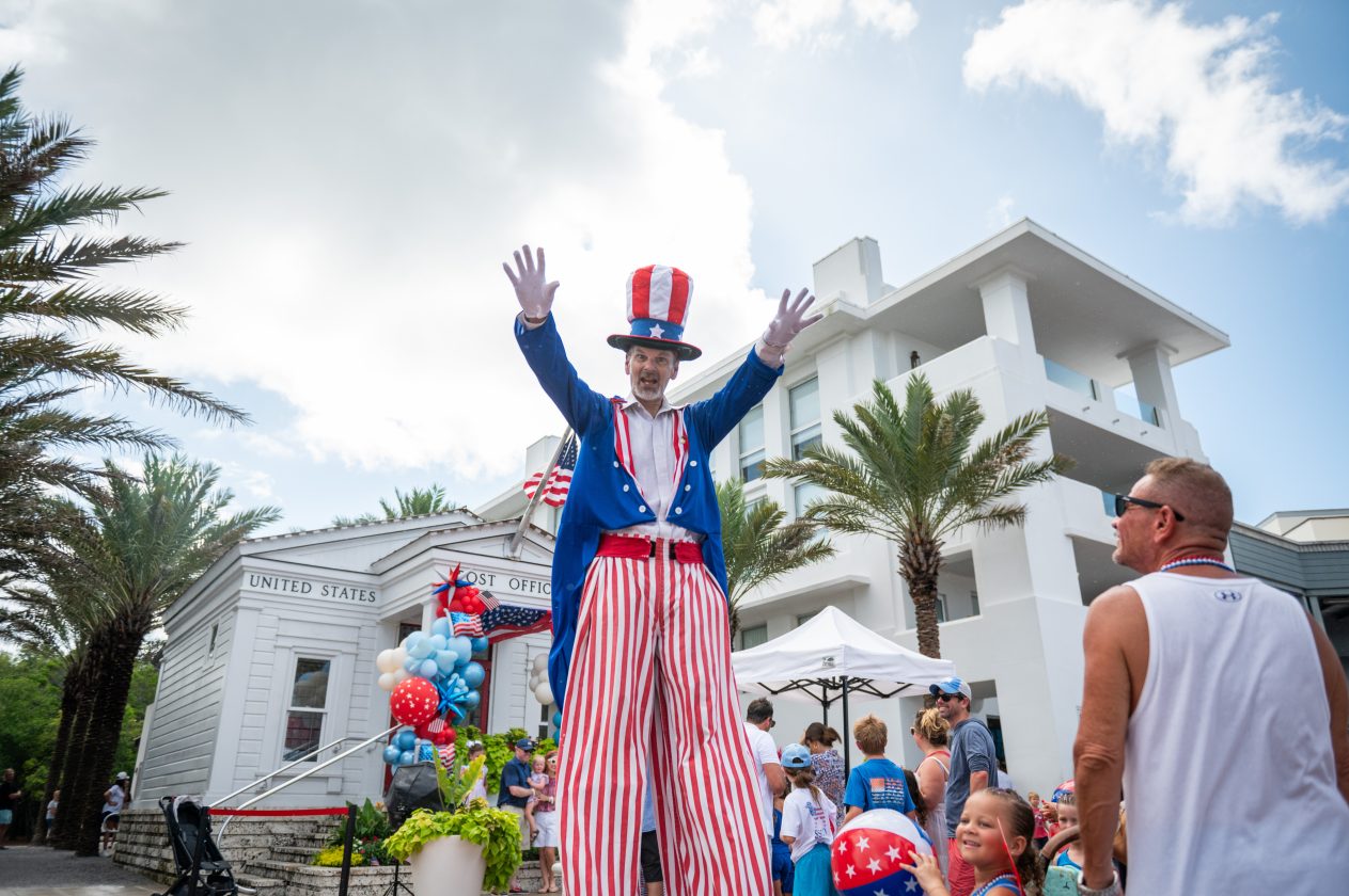Man dressed as Uncle Sam on stilts entertains crowd during patriotic celebration near white buildings and palm trees.