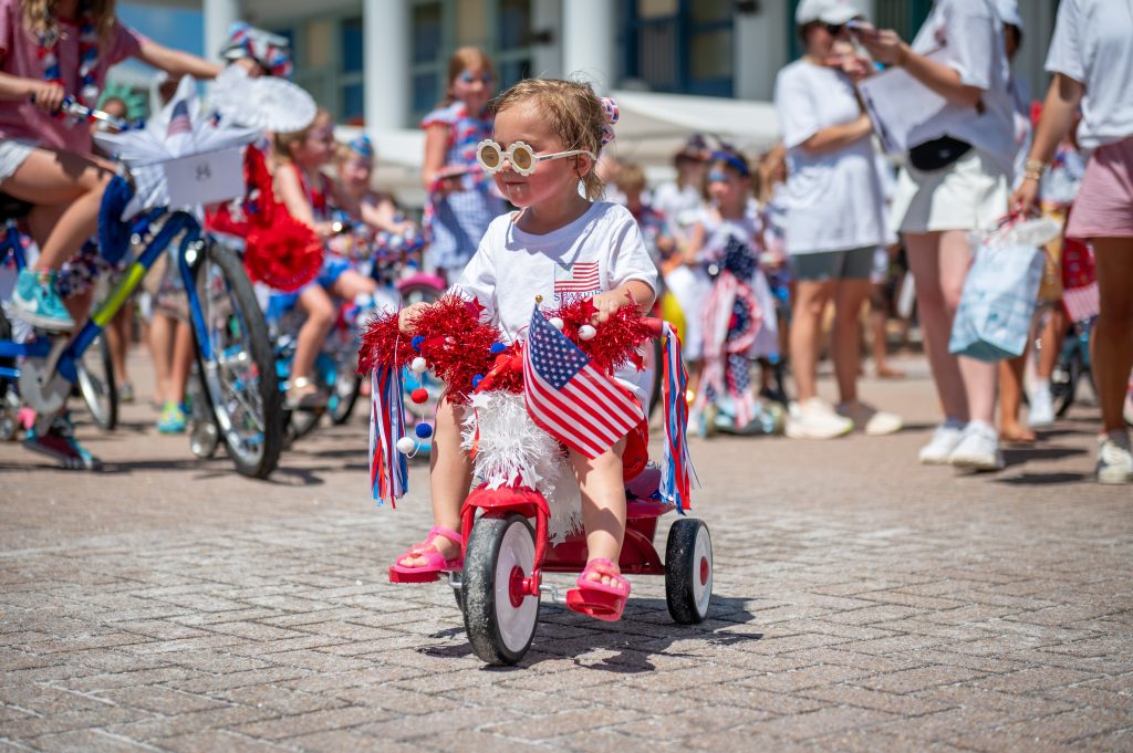 Toddler rides a tricycle decorated with red, white, and blue streamers and an American flag during a festive parade.