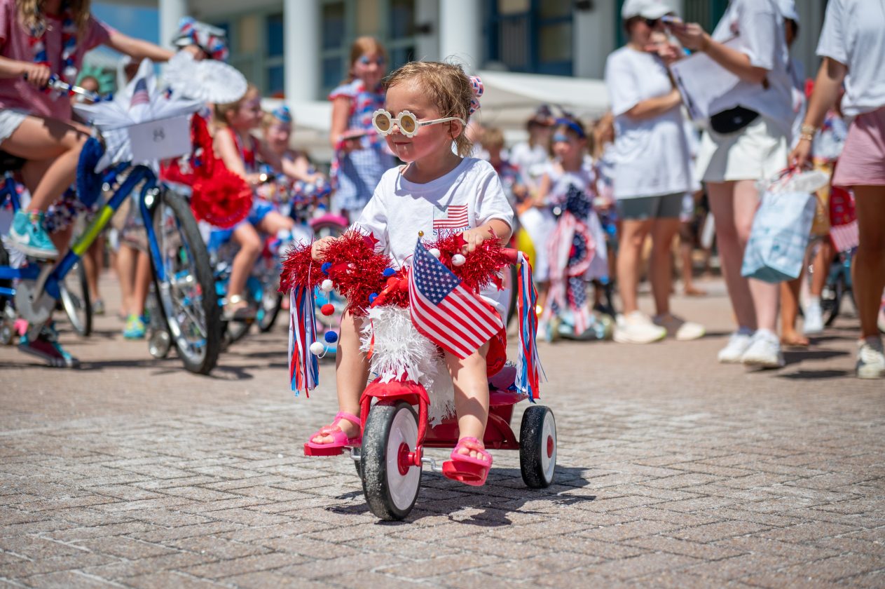 Toddler rides a decorated tricycle with an American flag during a festive parade with children and bikes.