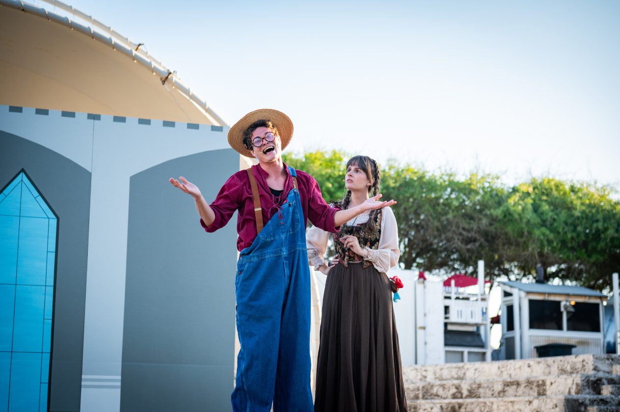 Two actors performing outdoors; one in overalls and hat, the other in brown dress.