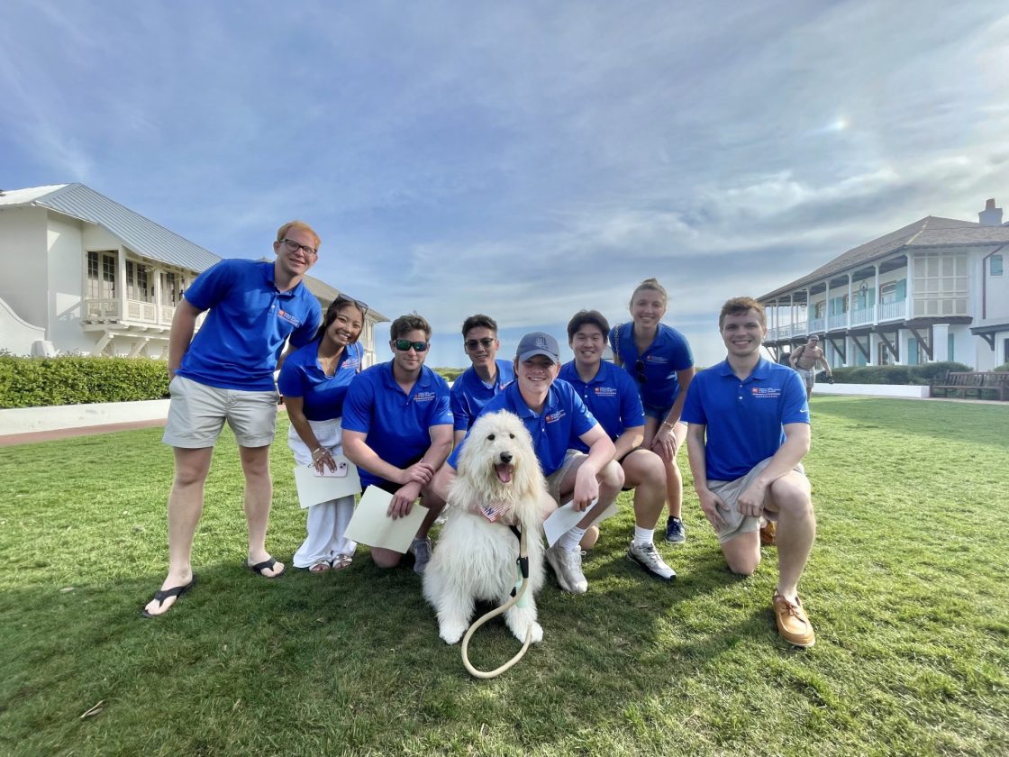 A group of people in matching blue shirts pose with a fluffy white dog on green lawn outdoors.