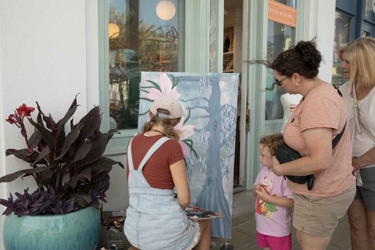 Artist painting floral artwork outdoors while a woman and child watch near storefront on sunny day.