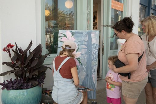 Artist painting floral artwork outdoors while a woman and child watch near storefront on sunny day.