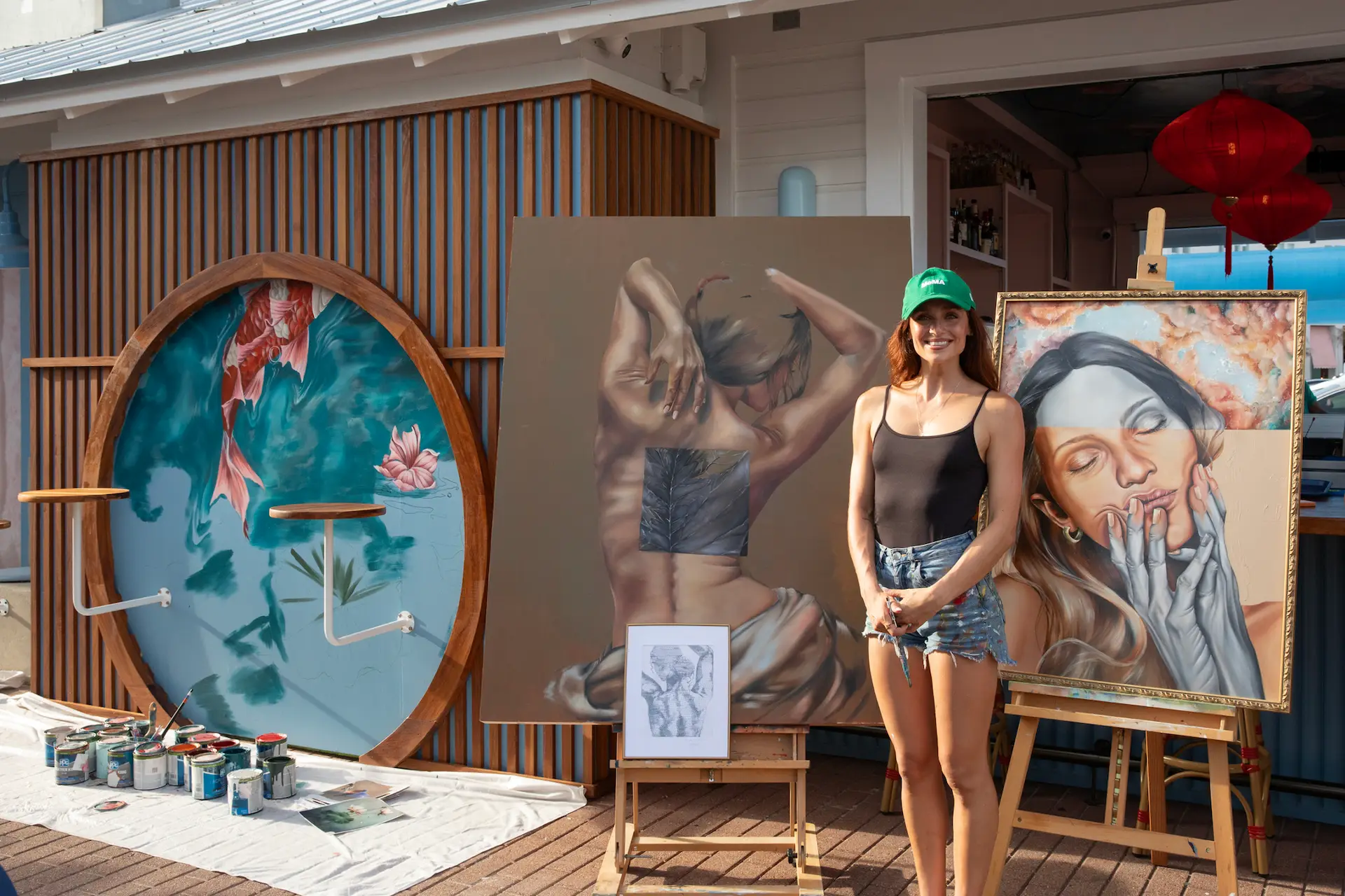 Artist standing between two large realistic paintings, outdoor art display with paint cans and brushes nearby.