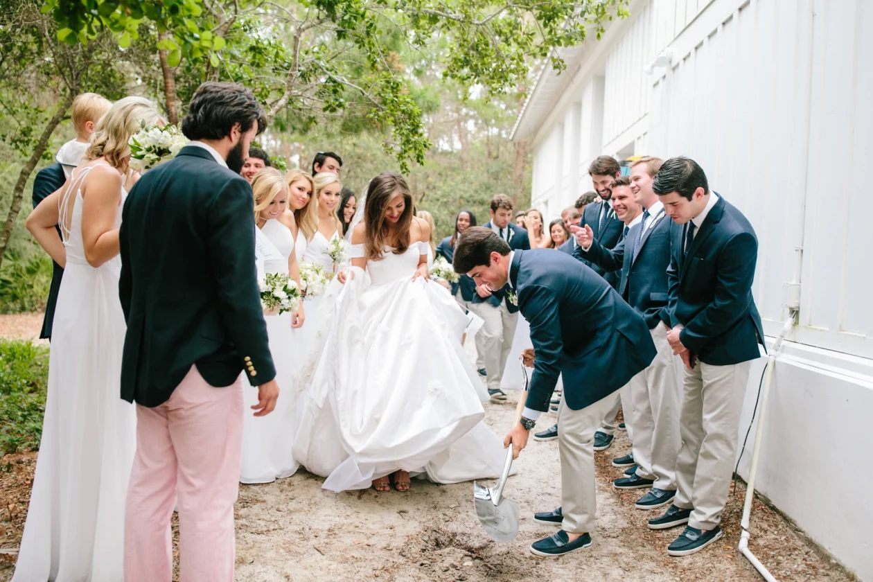 A bride surrounded by wedding party watches groom burying something, outdoor ceremony near building and trees.