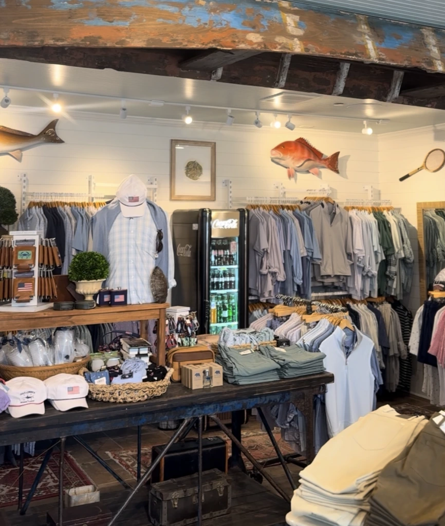 Coastal clothing boutique interior with folded shirts, hanging apparel, fishing decor, and a rustic ceiling.