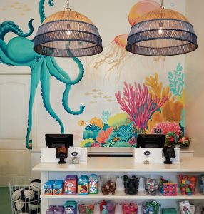Colorful undersea mural with octopus, coral, jellyfish behind store counter displaying treats, toys, and two registers.