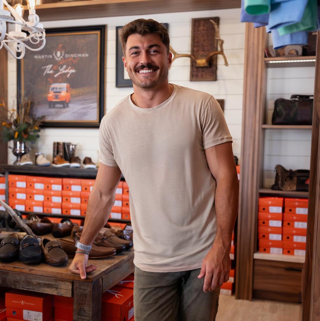 Smiling man in beige shirt poses inside a stylish shoe store with shelves of orange boxes.
