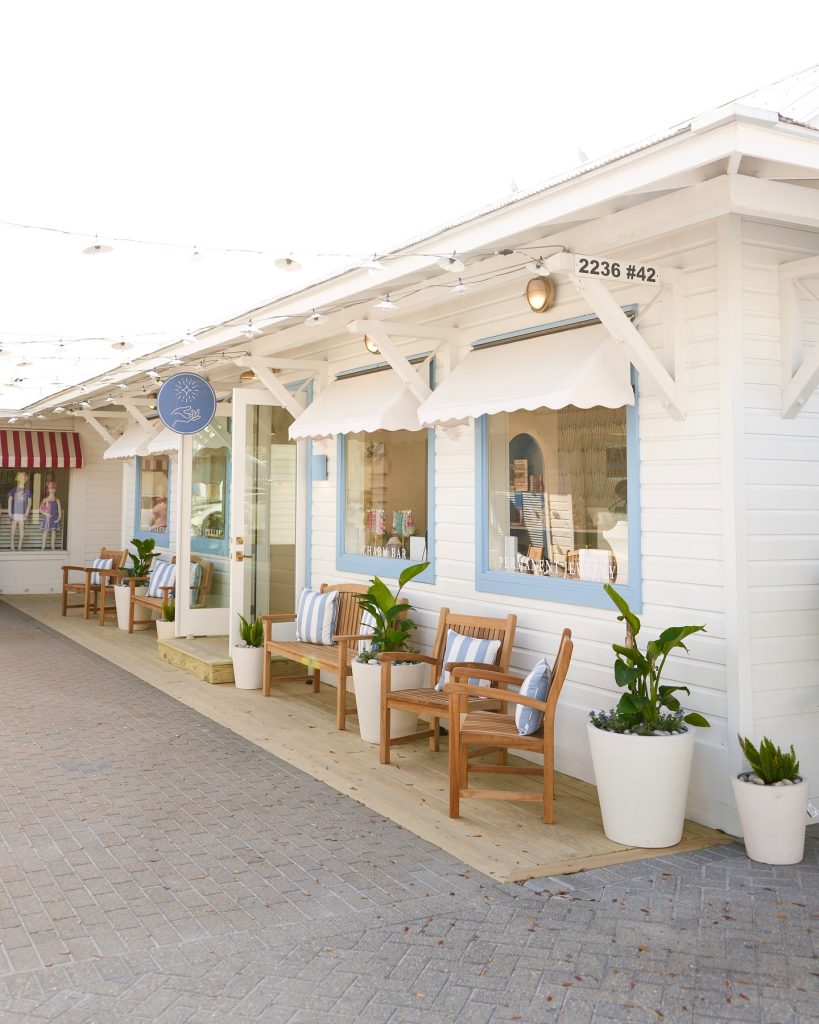 Bright seaside boutique with blue trim, wooden chairs, potted plants, and string lights overhead.