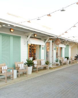 Cozy boutique storefront with cushioned wooden chairs, potted plants, mint green shutters, and string lights above.