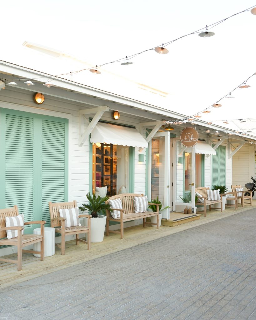 Cozy boutique storefront with cushioned wooden chairs, potted plants, mint green shutters, and string lights above.