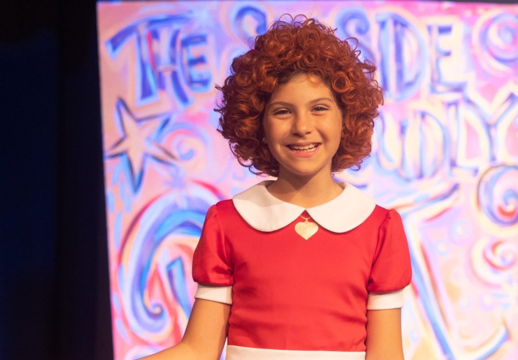 Smiling child actor in an Annie costume performing on a brightly lit stage.