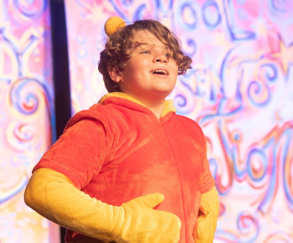 Child performer in a Winnie the Pooh costume singing on a colorful stage.