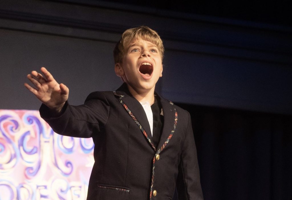 Young performer in blazer singing passionately onstage with one arm outstretched.